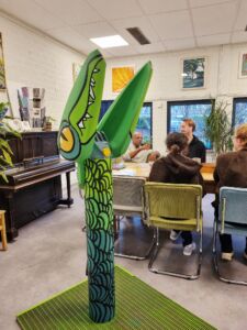 Een groene praatpaal, op de achtergrond mensen aan een tafel.