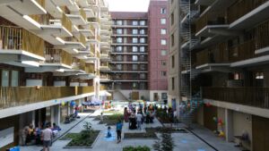 Je ziet de binnenplaats van gemengd wonen project Tango. Aan de randen grote nieuwbouwgebouwen en in het midden open ruimte met groen en mensen.