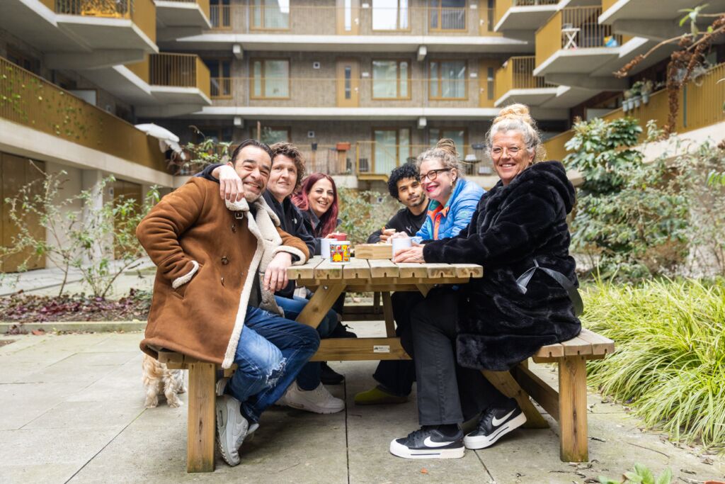 6 verschillende mensen zitten aan een picknicktafel met op de achtergrond een gebouw. Het ziet er gezellig uit, ze kijken vrolijk.